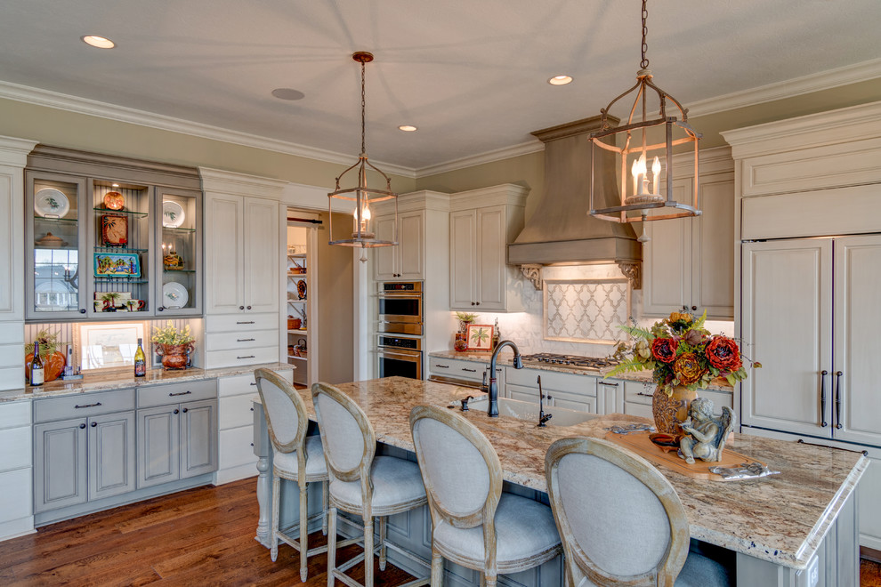 Design ideas for a large traditional l-shaped eat-in kitchen in Indianapolis with a farmhouse sink, raised-panel cabinets, white cabinets, panelled appliances, medium hardwood floors, with island, brown floor, beige benchtop, granite benchtops, white splashback and marble splashback.