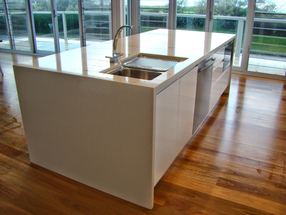 Photo of a mid-sized beach style galley kitchen in Sydney with an undermount sink, flat-panel cabinets, white cabinets, quartz benchtops, white splashback, glass sheet splashback, stainless steel appliances, dark hardwood floors and with island.