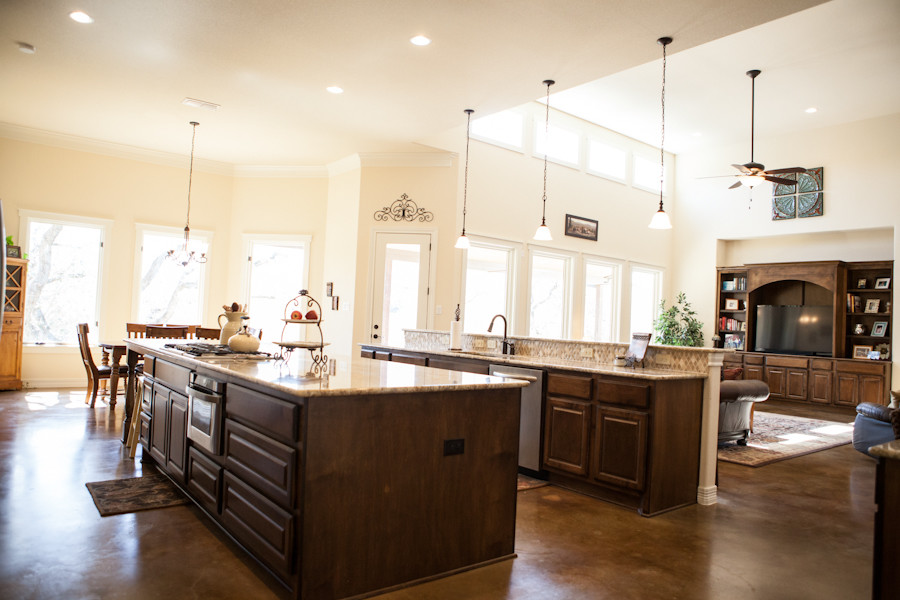 This is an example of a traditional eat-in kitchen in Austin with an undermount sink, raised-panel cabinets, brown cabinets, granite benchtops, beige splashback, travertine splashback, stainless steel appliances, concrete floors, multiple islands, brown floor and beige benchtop.
