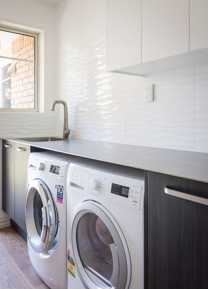 Inspiration for a small contemporary single-wall dedicated laundry room in Christchurch with an integrated sink, flat-panel cabinets, dark wood cabinets, stainless steel benchtops, white walls, medium hardwood floors, a side-by-side washer and dryer, brown floor and grey benchtop.