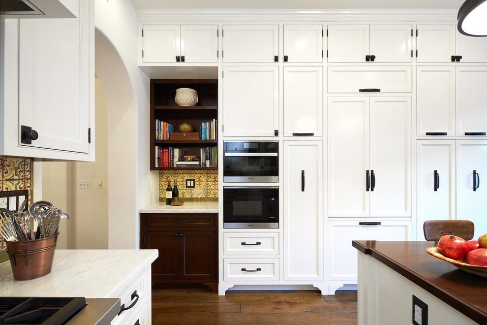 Photo of a mid-sized country l-shaped eat-in kitchen in Los Angeles with a farmhouse sink, glass-front cabinets, white cabinets, limestone benchtops, yellow splashback, mosaic tile splashback, black appliances, medium hardwood floors, with island and brown floor.