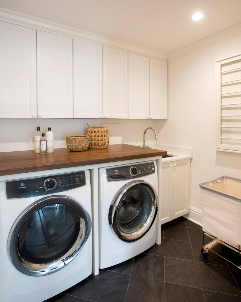 Exempel på en liten modern bruna brunt tvättstuga enbart för tvätt, med en undermonterad diskho, luckor med infälld panel, vita skåp, träbänkskiva, vita väggar, klinkergolv i keramik, en tvättmaskin och torktumlare bredvid varandra och grått golv