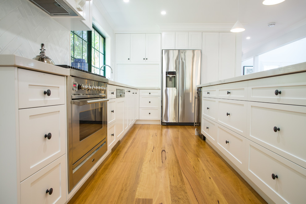 Photo of an expansive traditional galley eat-in kitchen in Brisbane with a single-bowl sink, shaker cabinets, white cabinets, solid surface benchtops, white splashback, subway tile splashback, stainless steel appliances, medium hardwood floors, with island, brown floor and white benchtop.