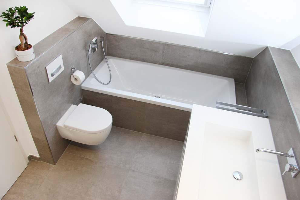 This is an example of a large contemporary bathroom in Munich with flat-panel cabinets, white cabinets, an alcove tub, a wall-mount toilet, gray tile, white walls, an integrated sink, solid surface benchtops, grey floor and white benchtops.