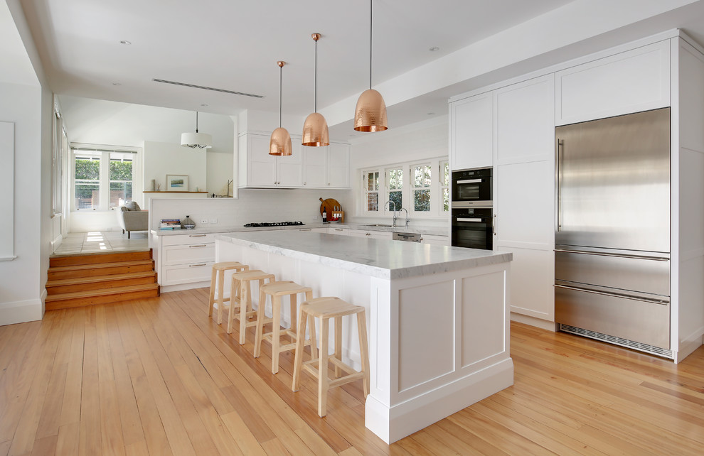 This is an example of a large transitional l-shaped eat-in kitchen in Sydney with a double-bowl sink, shaker cabinets, white cabinets, marble benchtops, white splashback, ceramic splashback, stainless steel appliances, light hardwood floors, with island and yellow floor.