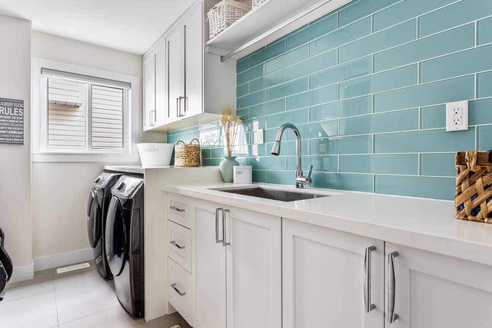 This is an example of a large beach style single-wall dedicated laundry room in Vancouver with an undermount sink, shaker cabinets, white cabinets, green splashback, glass tile splashback, white walls, ceramic floors, a side-by-side washer and dryer, white floor and white benchtop.