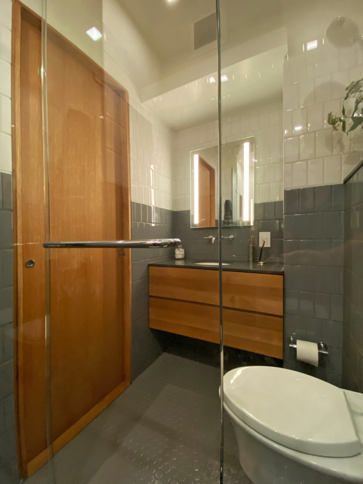 This is an example of a small contemporary master bathroom in Boston with flat-panel cabinets, medium wood cabinets, an alcove shower, a wall-mount toilet, gray tile, ceramic tile, white walls, ceramic floors, an undermount sink, soapstone benchtops, grey floor, a hinged shower door, black benchtops, a single vanity, a floating vanity, vaulted and decorative wall panelling.