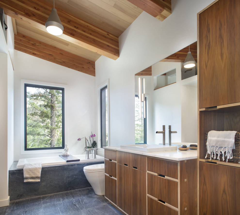 This is an example of a contemporary bathroom in Denver with flat-panel cabinets, medium wood cabinets, a drop-in tub, gray tile, white walls, a drop-in sink, white benchtops, a single vanity, a built-in vanity, exposed beam and vaulted.
