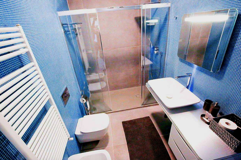 This is an example of a mid-sized modern 3/4 bathroom in Other with open cabinets, white cabinets, a curbless shower, a wall-mount toilet, blue tile, mosaic tile, white walls, porcelain floors, a vessel sink, stainless steel benchtops, grey floor, a sliding shower screen and grey benchtops.