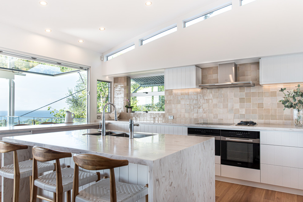 This is an example of a large beach style l-shaped eat-in kitchen in Sunshine Coast with a drop-in sink, white cabinets, beige splashback, porcelain splashback, black appliances, medium hardwood floors, with island and white benchtop.