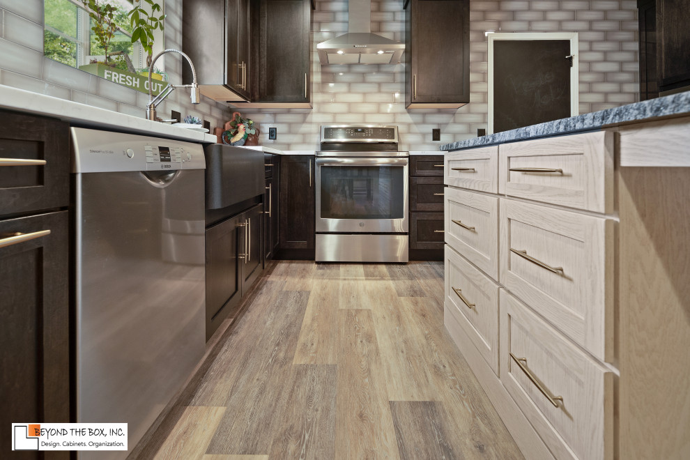 Example of a mid-sized transitional l-shaped medium tone wood floor and brown floor kitchen design with a farmhouse sink, shaker cabinets, black cabinets, granite countertops, gray backsplash, ceramic backsplash, stainless steel appliances, an island and white countertops