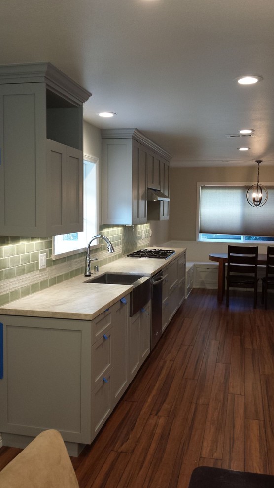 Example of a mid-sized transitional galley medium tone wood floor eat-in kitchen design with a farmhouse sink, shaker cabinets, white cabinets, granite countertops, green backsplash, glass tile backsplash, stainless steel appliances and no island
