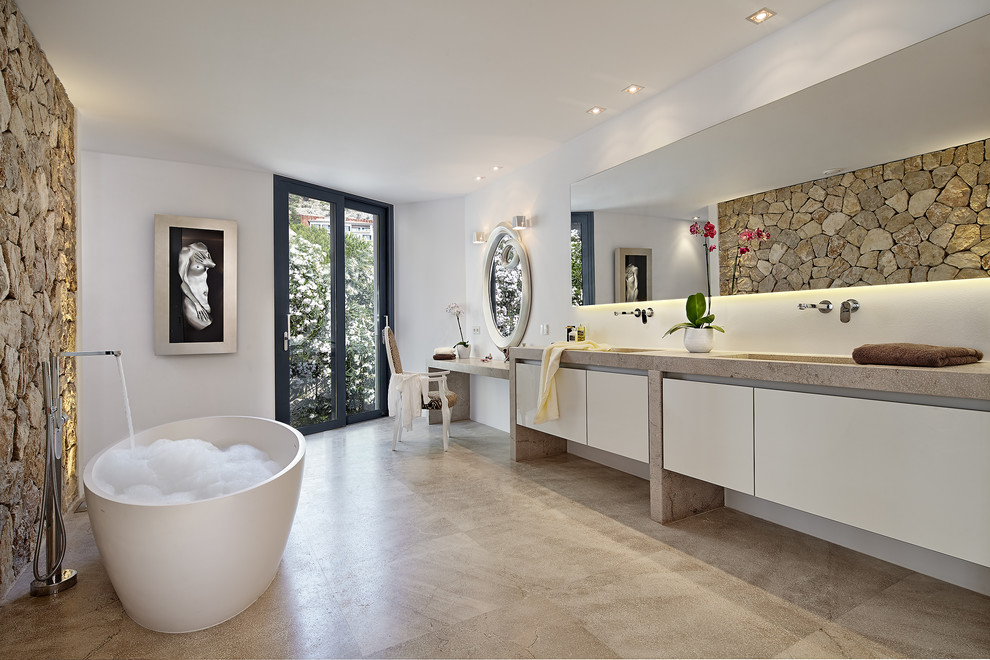 This is an example of a large contemporary master bathroom in Palma de Mallorca with a freestanding tub, white walls, limestone floors, an integrated sink, limestone benchtops, beige floor, beige benchtops, flat-panel cabinets, white cabinets, beige tile and stone tile.