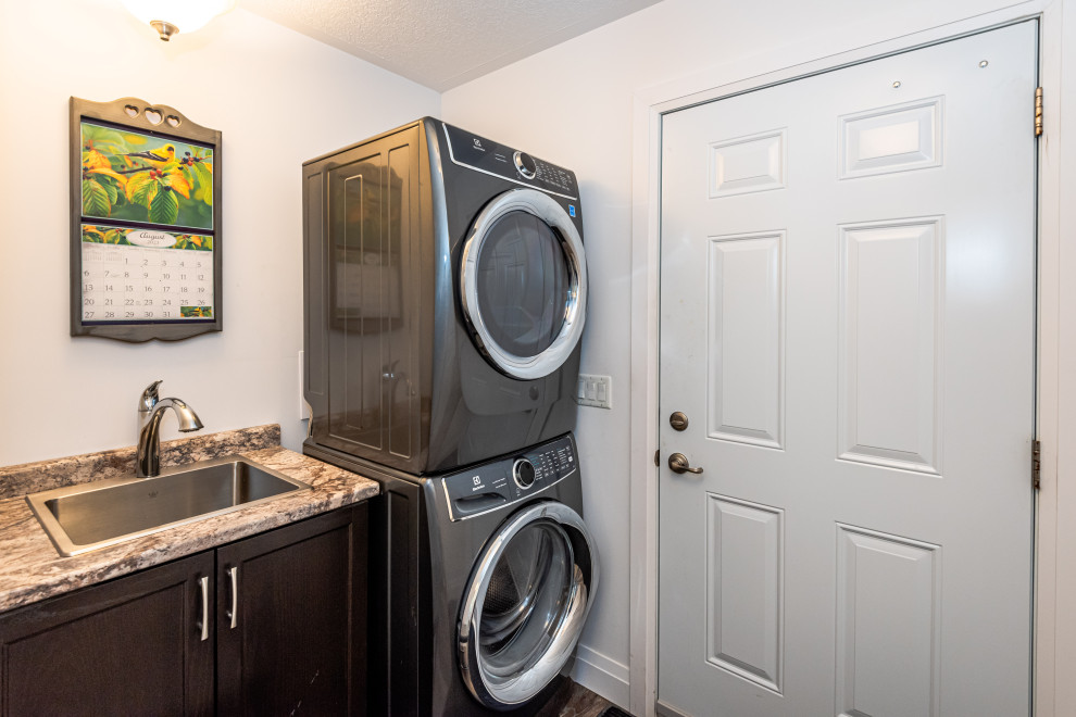 Photo of a single-wall dedicated laundry room in Toronto with a drop-in sink, recessed-panel cabinets, brown cabinets, white walls, vinyl floors, a stacked washer and dryer and brown floor.