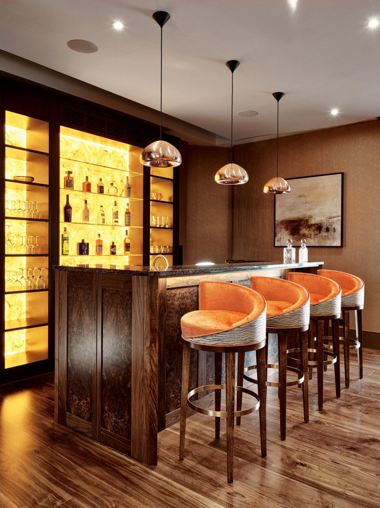 This is an example of a transitional galley seated home bar in London with shaker cabinets, medium wood cabinets, medium hardwood floors, brown floor and multi-coloured benchtop.