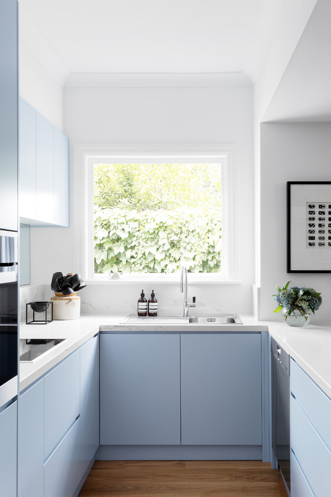 Photo of a contemporary u-shaped open plan kitchen in Melbourne with a drop-in sink, flat-panel cabinets, blue cabinets, metallic splashback, mirror splashback, stainless steel appliances, light hardwood floors and a peninsula.