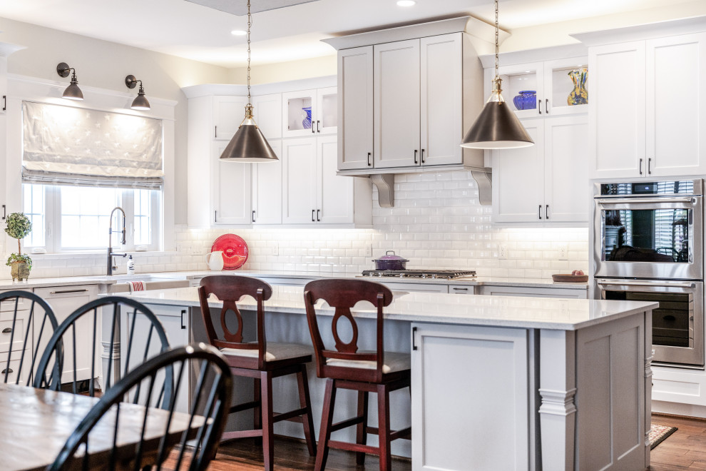 Example of a large farmhouse l-shaped medium tone wood floor, brown floor and wallpaper ceiling eat-in kitchen design in DC Metro with a farmhouse sink, shaker cabinets, white cabinets, quartz countertops, white backsplash, subway tile backsplash, stainless steel appliances, an island and white countertops