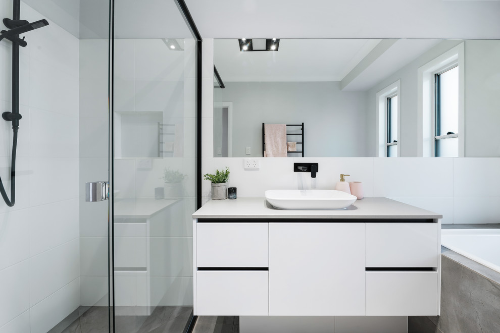 This is an example of a mid-sized contemporary kids bathroom in Other with flat-panel cabinets, white cabinets, a drop-in tub, a corner shower, a one-piece toilet, white tile, ceramic tile, white walls, a vessel sink, engineered quartz benchtops, grey floor, a hinged shower door, white benchtops and porcelain floors.