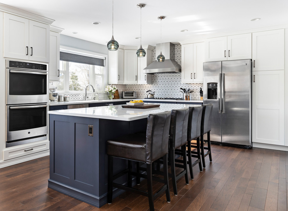 Design ideas for a large transitional l-shaped open plan kitchen in Detroit with blue cabinets, white splashback, ceramic splashback, stainless steel appliances, with island, white benchtop, shaker cabinets, medium hardwood floors and brown floor.