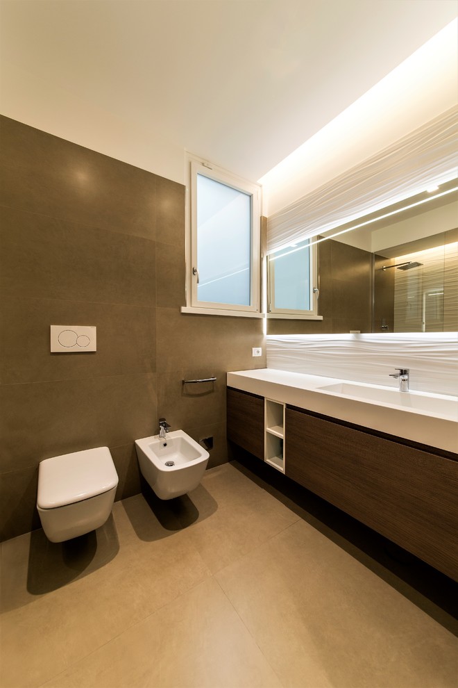 This is an example of a mid-sized contemporary master bathroom in Rome with beaded inset cabinets, dark wood cabinets, a drop-in tub, a shower/bathtub combo, a one-piece toilet, brown tile, porcelain tile, brown walls, porcelain floors, an integrated sink, solid surface benchtops, beige floor, a hinged shower door and white benchtops.