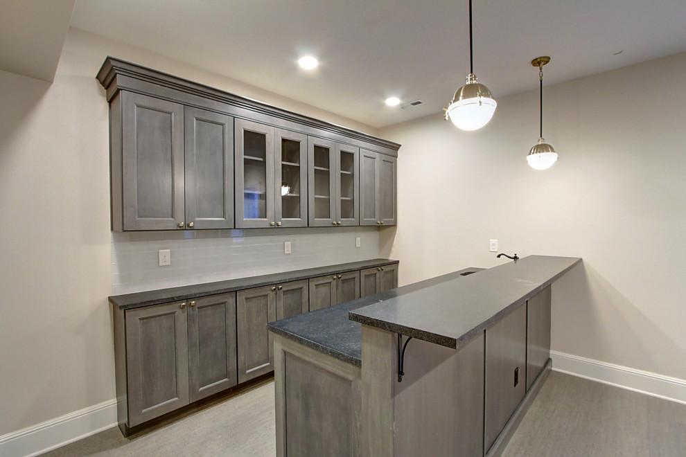 Photo of a small transitional galley wet bar in Denver with an undermount sink, shaker cabinets, grey cabinets, laminate benchtops, white splashback, glass tile splashback, laminate floors, brown floor and black benchtop.
