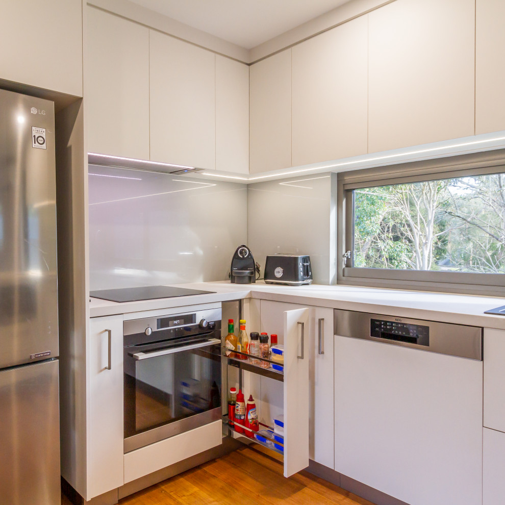 Inspiration for a mid-sized contemporary u-shaped eat-in kitchen in Melbourne with a drop-in sink, flat-panel cabinets, white cabinets, quartz benchtops, white splashback, glass sheet splashback, stainless steel appliances, medium hardwood floors, a peninsula, brown floor and white benchtop.