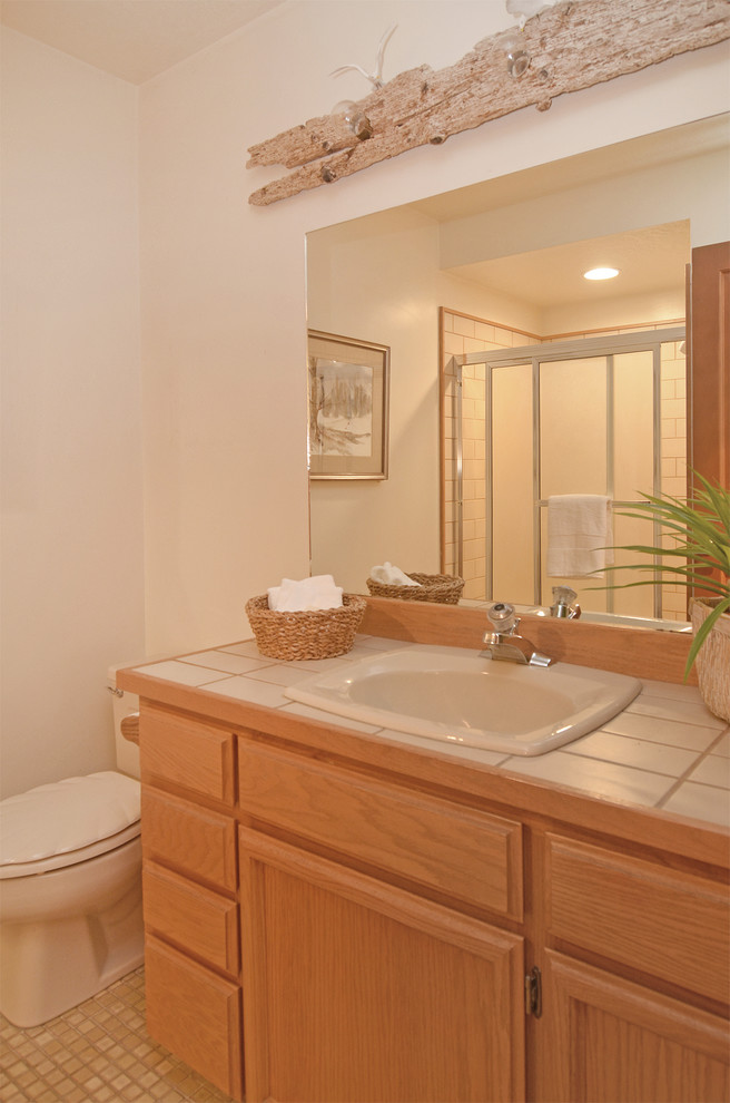 Photo of a small contemporary bathroom in Seattle with a drop-in sink, recessed-panel cabinets, medium wood cabinets, tile benchtops, an alcove shower, white tile, ceramic tile, beige walls and vinyl floors.