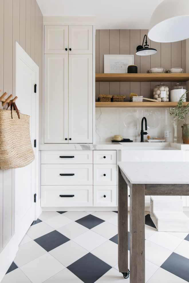 Large transitional galley utility room in Phoenix with a farmhouse sink, recessed-panel cabinets, white cabinets, marble benchtops, grey splashback, marble splashback, white walls, ceramic floors, a stacked washer and dryer, black floor, white benchtop and planked wall panelling.