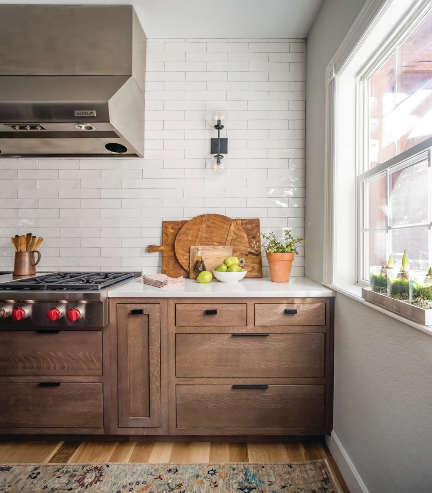Inspiration for a mid-sized country l-shaped kitchen in Denver with a farmhouse sink, shaker cabinets, quartz benchtops, white splashback, subway tile splashback, stainless steel appliances, light hardwood floors, with island, brown floor and white benchtop.