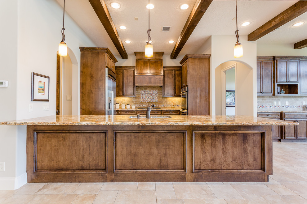 Inspiration for a large mediterranean u-shaped open plan kitchen in Houston with a drop-in sink, recessed-panel cabinets, brown cabinets, granite benchtops, beige splashback, stone tile splashback, stainless steel appliances, travertine floors, with island, beige floor and beige benchtop.