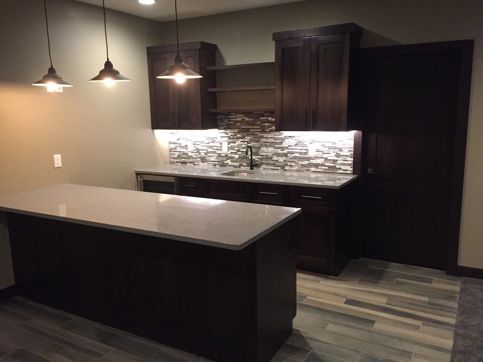 Mid-sized arts and crafts galley seated home bar in Cedar Rapids with an undermount sink, shaker cabinets, dark wood cabinets, quartz benchtops, multi-coloured splashback, matchstick tile splashback, medium hardwood floors, brown floor and grey benchtop.