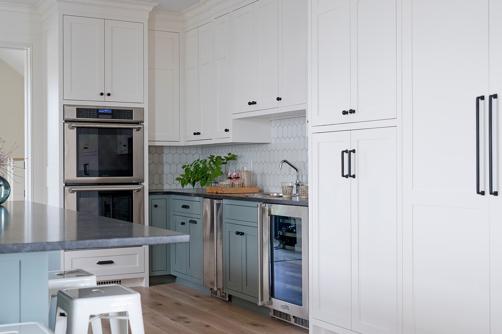 Photo of a large beach style l-shaped kitchen in Portland Maine with shaker cabinets, blue cabinets, granite benchtops, white splashback, porcelain splashback, stainless steel appliances, light hardwood floors, with island, beige floor and black benchtop.