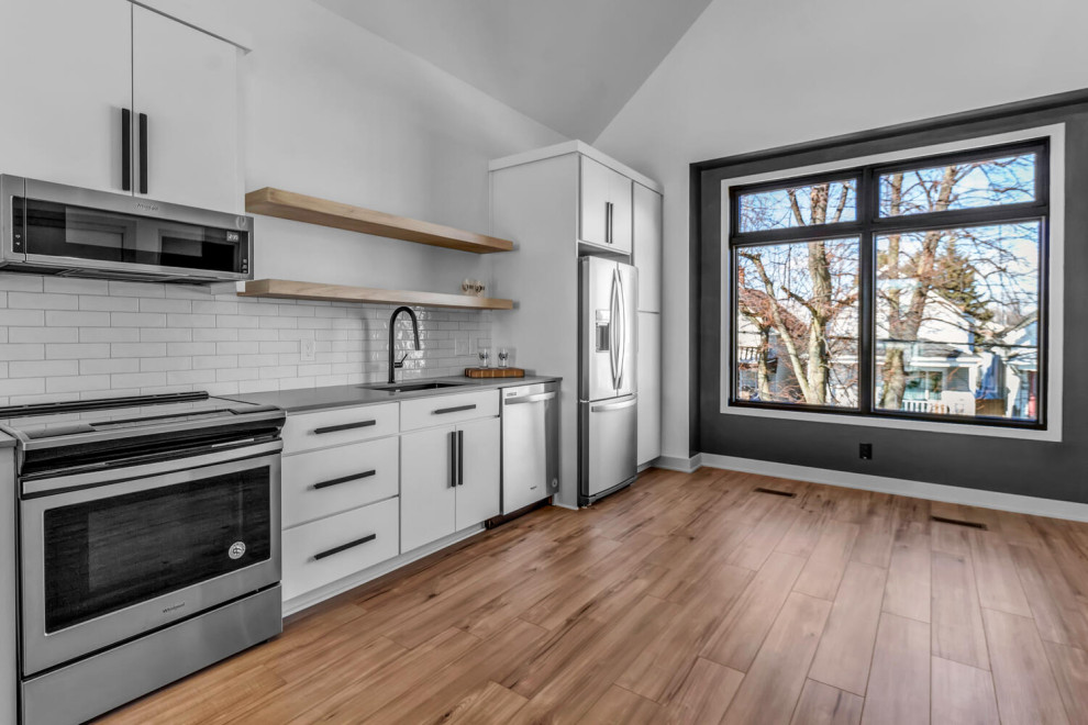 This is an example of a mid-sized modern single-wall eat-in kitchen in New York with an undermount sink, flat-panel cabinets, white cabinets, quartz benchtops, white splashback, subway tile splashback, stainless steel appliances, light hardwood floors, a peninsula, beige floor and grey benchtop.