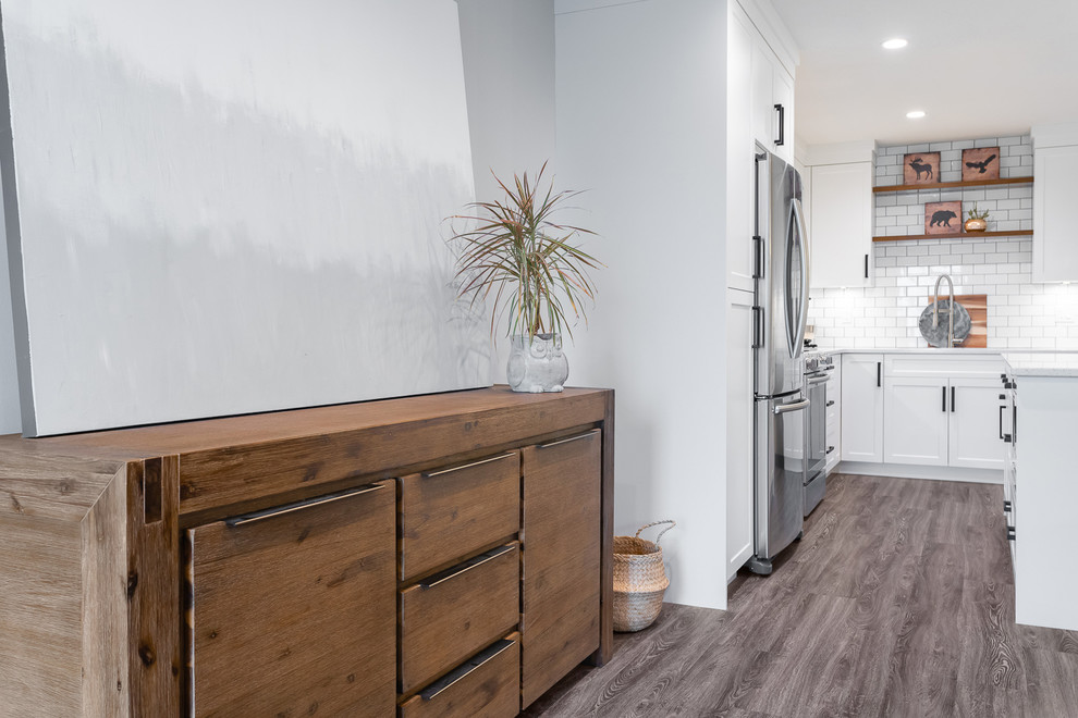 Inspiration for a mid-sized transitional l-shaped brown floor and vinyl floor eat-in kitchen remodel in Vancouver with an undermount sink, white cabinets, quartz countertops, white backsplash, subway tile backsplash, stainless steel appliances, an island, white countertops and shaker cabinets