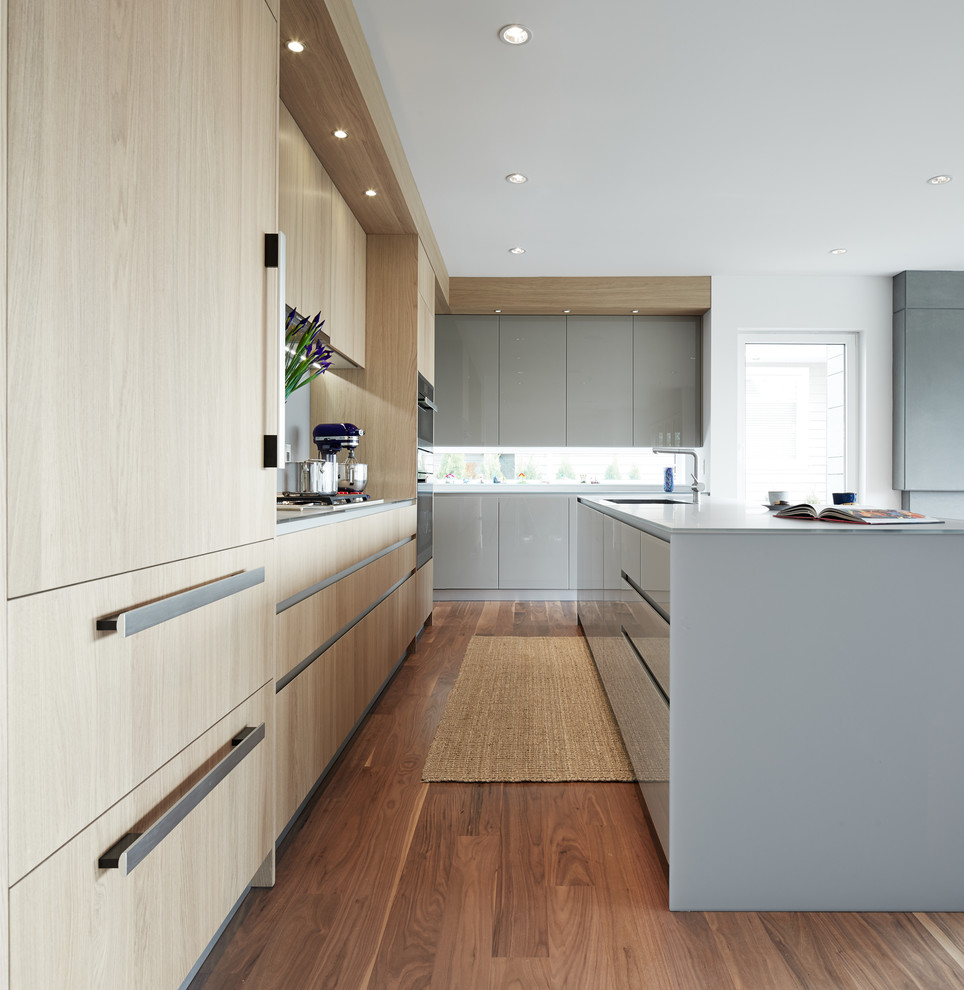 Mid-sized contemporary l-shaped open plan kitchen in Vancouver with an undermount sink, flat-panel cabinets, light wood cabinets, dark hardwood floors, with island, glass benchtops, grey splashback and stainless steel appliances.