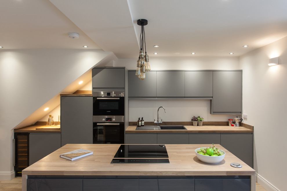 Contemporary galley kitchen in London with a drop-in sink, flat-panel cabinets, grey cabinets, wood benchtops, black appliances, light hardwood floors, with island, beige floor and beige benchtop.
