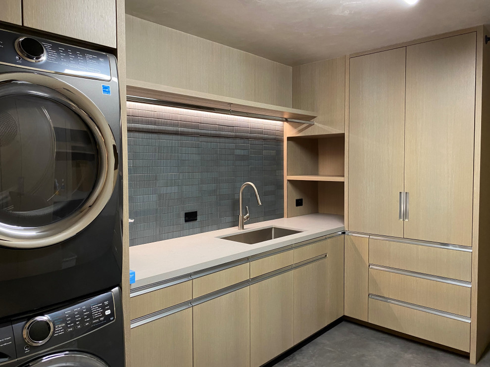 Mid-sized contemporary l-shaped dedicated laundry room in Other with a single-bowl sink, flat-panel cabinets, light wood cabinets, grey splashback, stone tile splashback, concrete floors, a stacked washer and dryer, grey floor, white benchtop and wood walls.