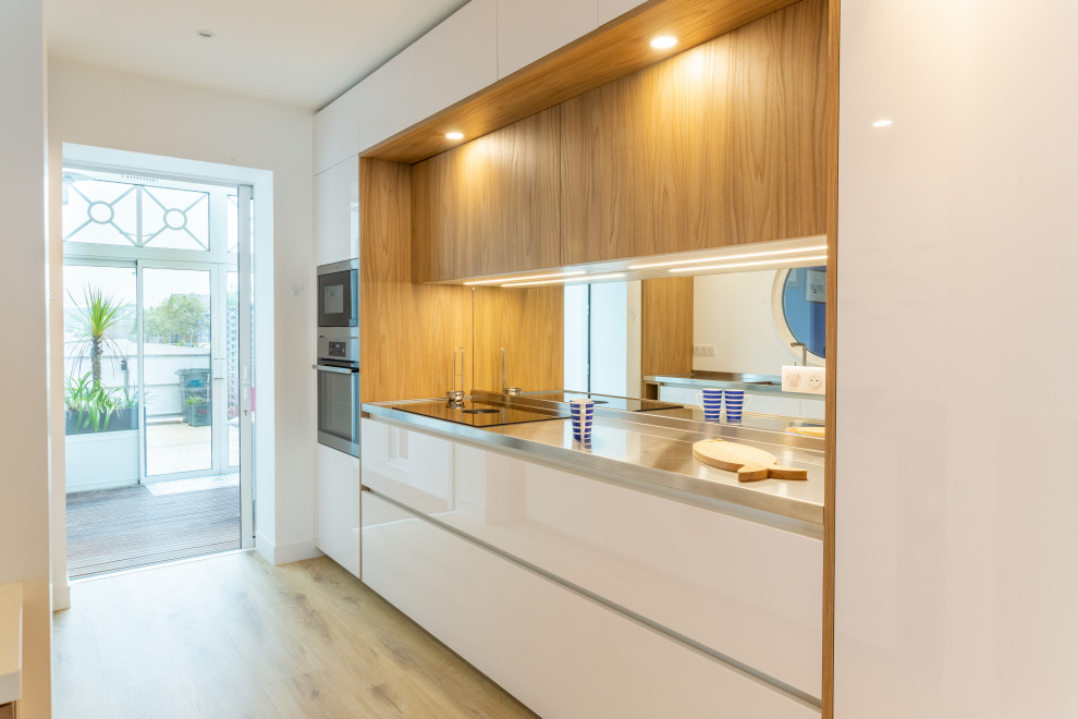 Foto på ett litet maritimt kök, med en integrerad diskho, luckor med profilerade fronter, vita skåp, bänkskiva i rostfritt stål, spegel som stänkskydd, rostfria vitvaror och ljust trägolv