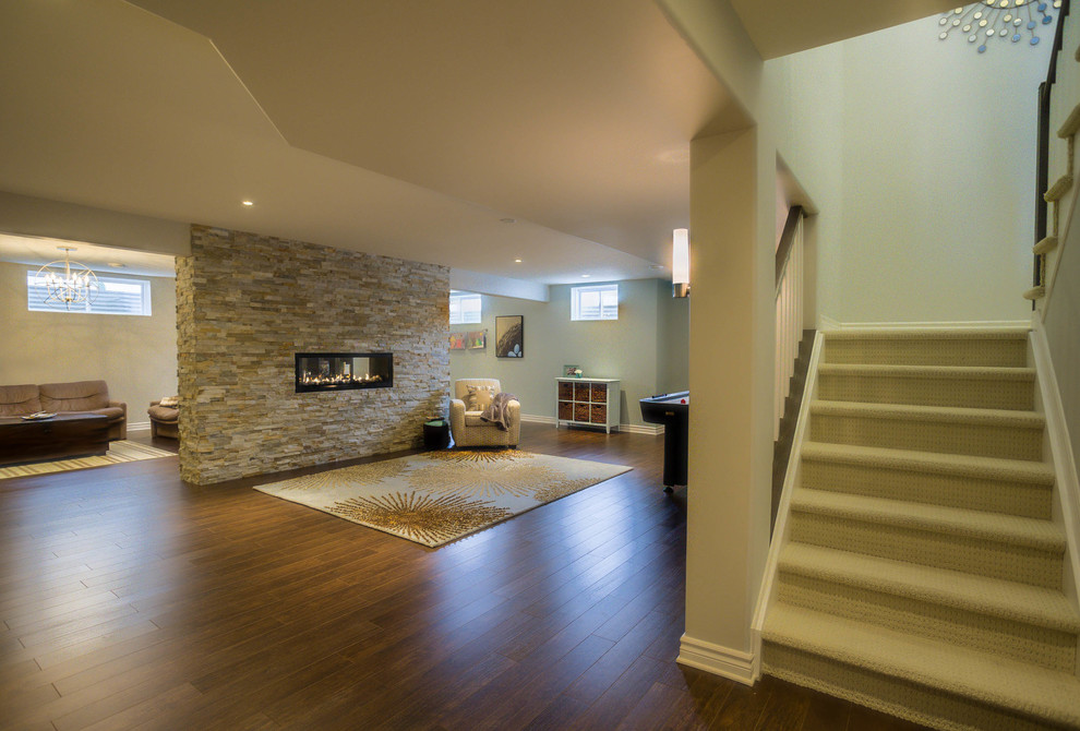 Large transitional fully buried basement in Ottawa with laminate floors, a two-sided fireplace and a stone fireplace surround.