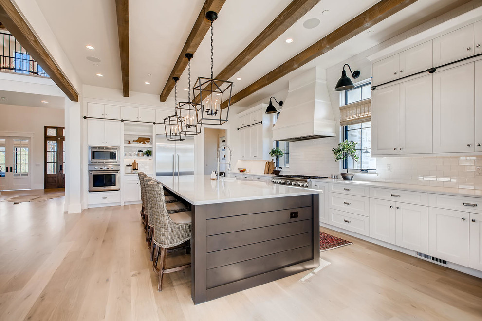Photo of a large country kitchen in Denver with a farmhouse sink, shaker cabinets, white cabinets, quartz benchtops, white splashback, subway tile splashback, stainless steel appliances, light hardwood floors, with island, white floor and white benchtop.