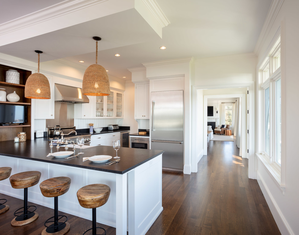 Design ideas for a large beach style l-shaped kitchen in Providence with a farmhouse sink, recessed-panel cabinets, white cabinets, granite benchtops, white splashback, timber splashback, stainless steel appliances, dark hardwood floors, a peninsula, brown floor and black benchtop.