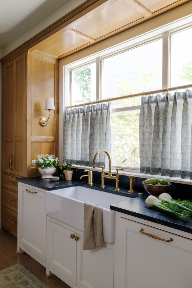 This is an example of a mid-sized traditional laundry room in Houston with a farmhouse sink, shaker cabinets, white cabinets, soapstone benchtops, multi-coloured splashback, ceramic splashback, white walls, light hardwood floors, brown floor and black benchtop.