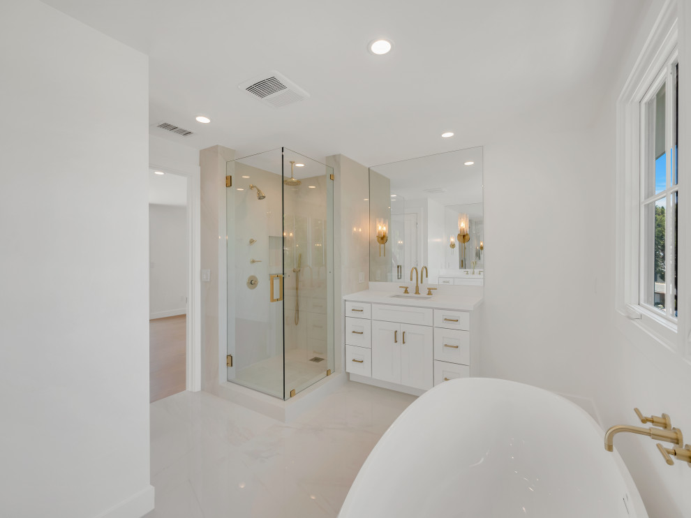 На фото: большая главная ванная комната в стиле кантри с фасадами в стиле шейкер, белыми фасадами, отдельно стоящей ванной, угловым душем, раздельным унитазом, белой плиткой, керамогранитной плиткой, белыми стенами, мраморным полом, врезной раковиной, столешницей из известняка, белым полом, душем с распашными дверями, белой столешницей, сиденьем для душа, тумбой под одну раковину и встроенной тумбой с