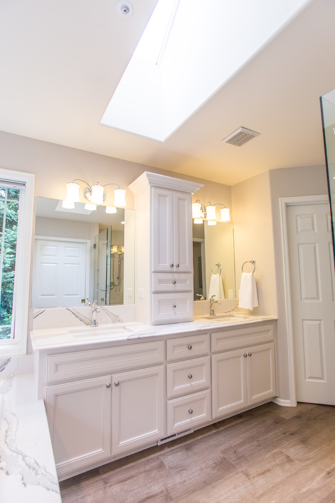 Inspiration for a mid-sized traditional master bathroom in Seattle with beaded inset cabinets, beige cabinets, a corner tub, a corner shower, white tile, stone slab, grey walls, light hardwood floors, an undermount sink, quartzite benchtops, grey floor, a hinged shower door and white benchtops.