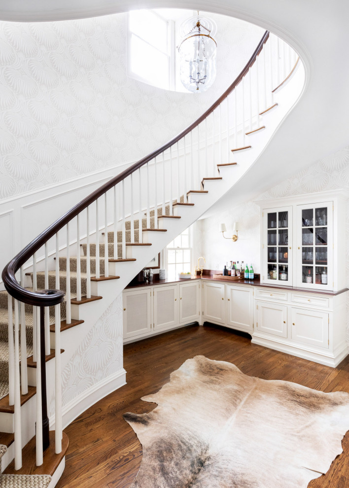 На фото: большая изогнутая деревянная лестница в стиле неоклассика (современная классика) с ступенями с ковровым покрытием, любыми перилами и обоями на стенах с