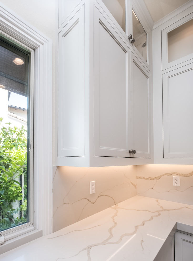 Enclosed kitchen - mid-sized transitional l-shaped porcelain tile and beige floor enclosed kitchen idea in Orange County with a farmhouse sink, recessed-panel cabinets, white cabinets, quartz countertops, white backsplash, marble backsplash, stainless steel appliances and an island