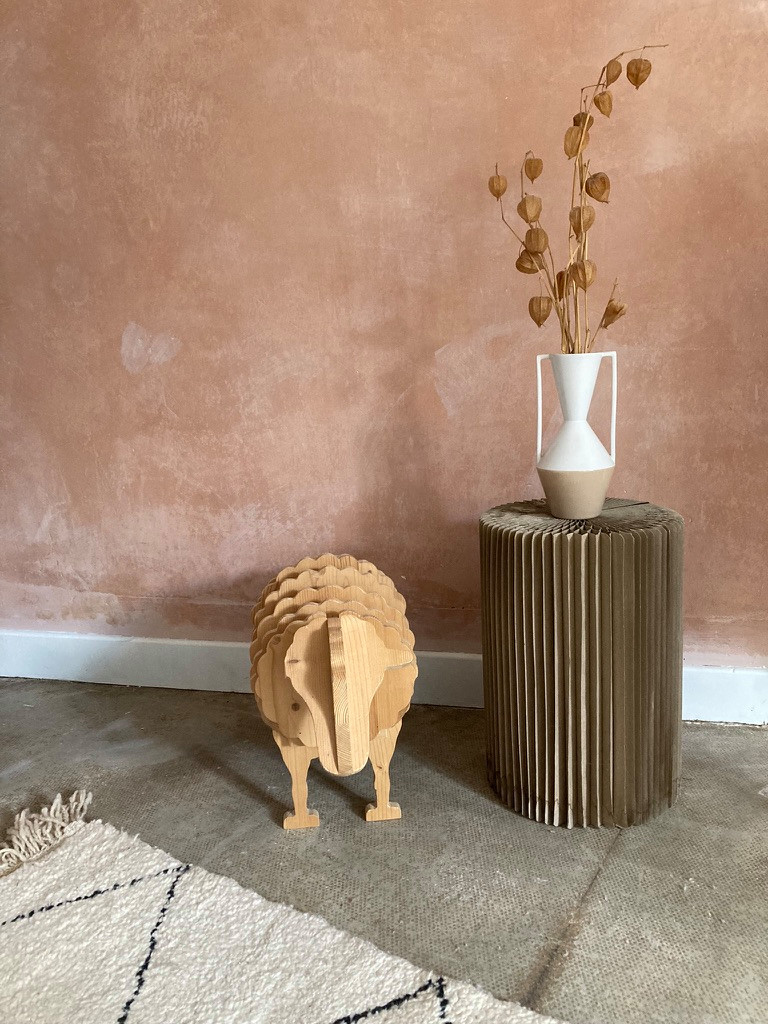 Détail du salon de télévision : un tabouret pliable en carton, un vase artisanal, un bouquet de fleurs séchées, un mouton en bois et un tapis berbère.