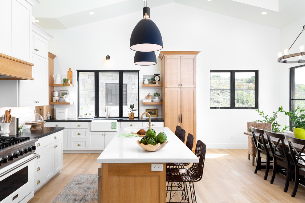 Large country eat-in kitchen in Salt Lake City with a farmhouse sink, shaker cabinets, light wood cabinets, solid surface benchtops, white splashback, ceramic splashback, stainless steel appliances, light hardwood floors, multiple islands, beige floor and black benchtop.