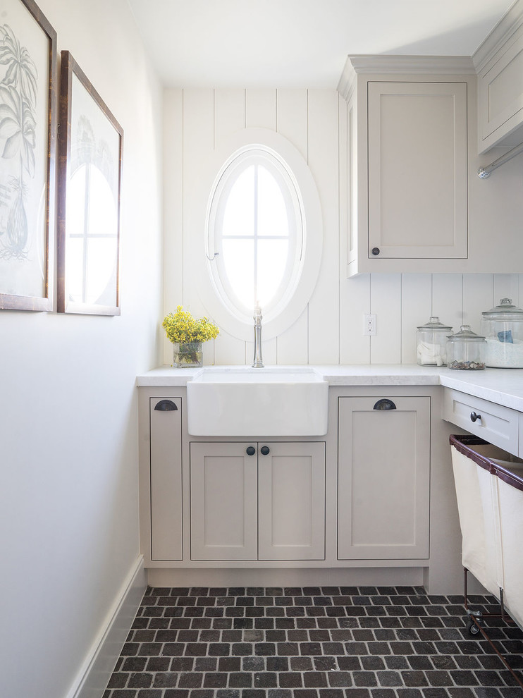 Photo of a beach style l-shaped dedicated laundry room in Orange County with a farmhouse sink, shaker cabinets, grey cabinets, white walls, brown floor and white benchtop.