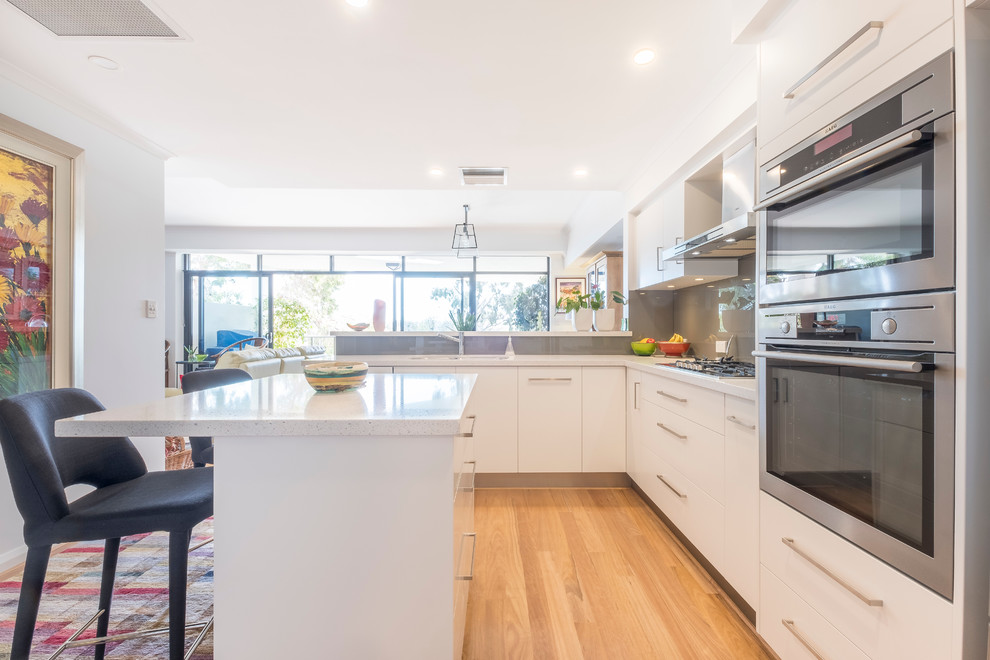 Photo of a contemporary kitchen in Perth with an undermount sink, flat-panel cabinets, white cabinets, quartz benchtops, grey splashback, glass sheet splashback, stainless steel appliances, with island, white benchtop and light hardwood floors.